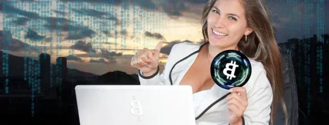 A person sitting behind a laptop holding a Bitcoin coin. The person is smiling and giving a thumbs up.